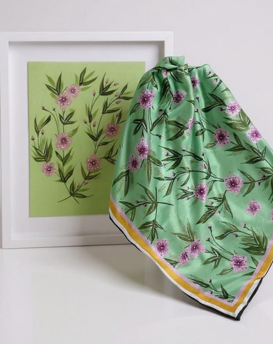 Green floral scarf with pink flowers and yellow border on a white surface, next to a framed artwork with green background and pink flowers.
